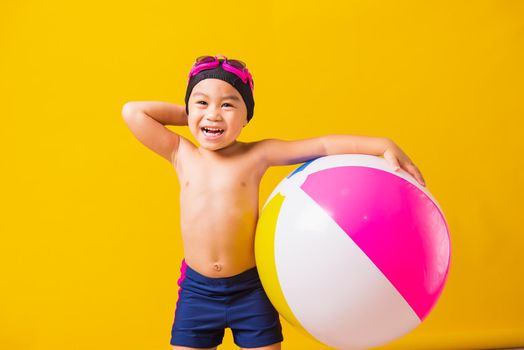 Summer vacation concept, Portrait Asian happy cute little child boy smiling in swimsuit hold beach ball, Kid having fun with inflatable ball in summer vacation, studio shot isolated yellow background