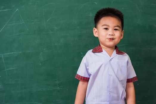 Back to School. Happy Asian funny cute little child boy from kindergarten in student uniform smiling on green school blackboard, First time to school education concept, studio shot