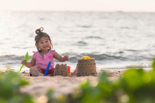 Happy fun Asian child cute little girl playing sand with toy sand tools at a tropical sea beach in holiday summer on sunset time, tourist trip concept