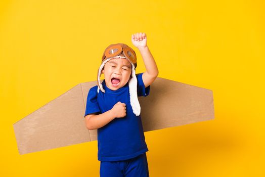 Happy Asian handsome funny child or kid little boy smile wear pilot hat play and goggles raise hand up with toy cardboard airplane wings flying, studio shot isolated yellow background, Startup freedom