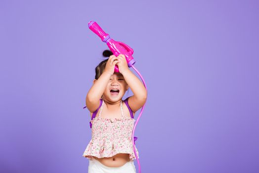 Thai child funny hold toy water pistol and smile, Happy Asian little girl holding plastic water gun, studio shot isolated on purple background, Thailand Songkran festival day national culture concept