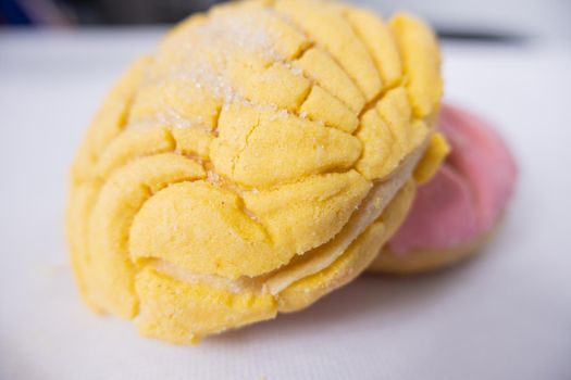 Two pieces of Mexican sweet bread known as concha on white table. Pice of yellow sweet bread leaning against pink piece of bread above table. Traditional Mexican desserts