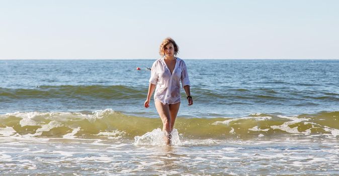 young blonde woman in a wet white shirt coming out of the water near the seashore on sunny summer day