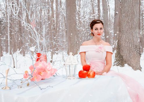 young beautiful woman in a pink dress sits at a table in the forest on a winter day