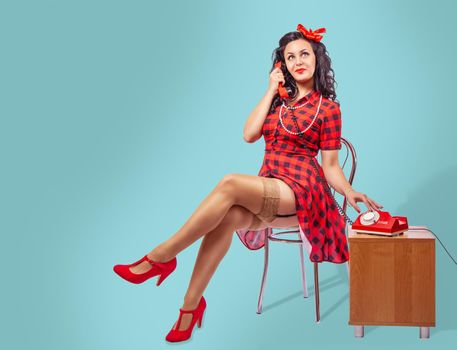 happy young pinup woman in red dress and nylon stockings sitting on a chair and talking on phone over green background