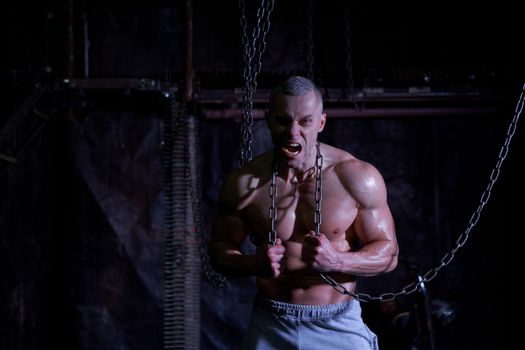 Primal instinct. Angry muscular man screaming at camera and breaking chains on chest, dark background