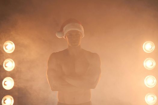 Portrait of muscular man wearing Christmas Santa hat, folded hands and smile on smoky background Macho Shirtless Naked torso stripper Hot sexy Santa guy Seductive male for holiday