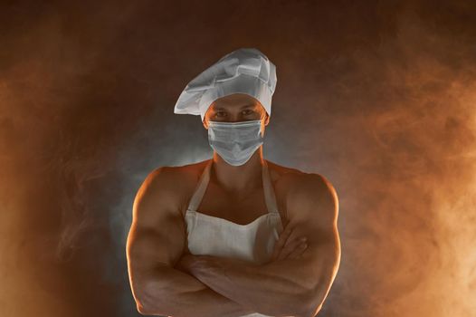 New normal concept. Portrait of muscular chef wearing protective medical mask white apron and chef hat, posing with folded arms on smoky background male housewife. husband in kitchen. brutal butcher.