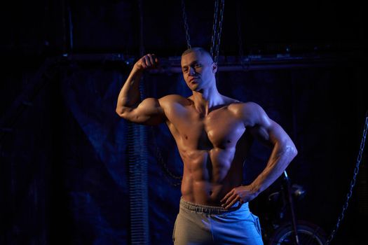 Young shirtless muscular man standing among metal chains, looking seriously at camera, copy space with his back to the camera shows biceps