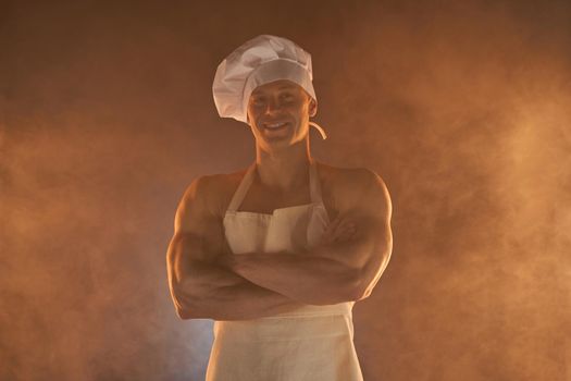 Portrait of muscular chef wearing white apron and chef hat, posing with folded arms on smoky background male housewife. husband in kitchen. brutal butcher.
