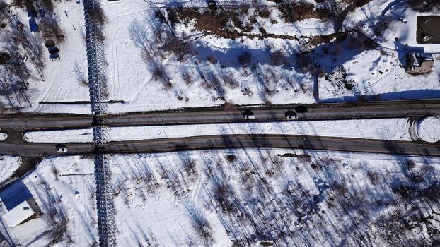 View from the height of the winter road. Along the road there are trees and mountainous terrain. Passing cars, green bus. The shadow of the trees falls on the snow. The house is visible.