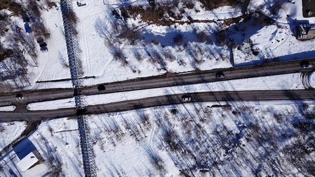 View from the height of the winter road. Along the road there are trees and mountainous terrain. Passing cars, green bus. The shadow of the trees falls on the snow. The house is visible.