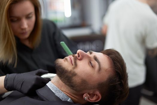 Young adult beautiful caucasian woman hairdresser cuts beard handsome man at modern barbershop Happy gay sitting chair men beauty salon. beard care. metrosexual. Female barber in black gloves serving