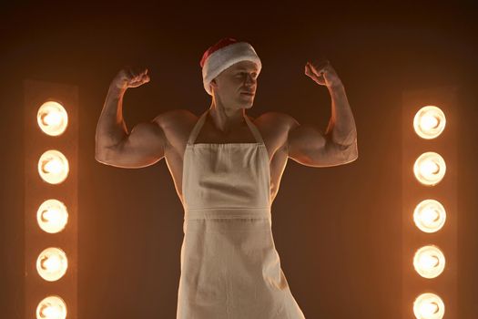 Hot Christmas dessert Sexy muscular Chef wearing apron and Santa hat show biceps smiling, lamps illumination and smoke on background cuisine. male housewife. husband in kitchen. brutal butcher.