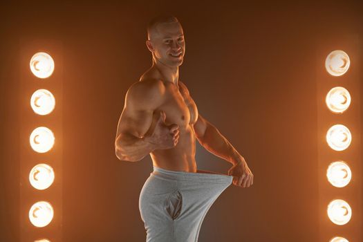Great male size. Young muscular guy pulling back his pants and showing thumb up gesture, being proud and satisfied with his genitals, lamps illumination on background Weight loss concept Diet