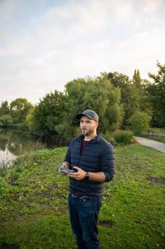 Concentrated man standing next to lake and holding remote control. Man in park with trees and boats on lake as background. People interacting with technology outside