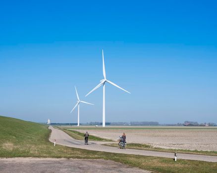 zierikzee, netherlands, 30 march 2021: people on bicycle and wind turbines in rural landscape of schouwen duiveland in dutch province of zeeland under blue sky