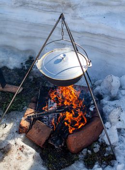 Over the fire hangs a pot in which to cook food. On a hook on a tripod, steam comes out of the pan. Winter Camping outdoor cooking