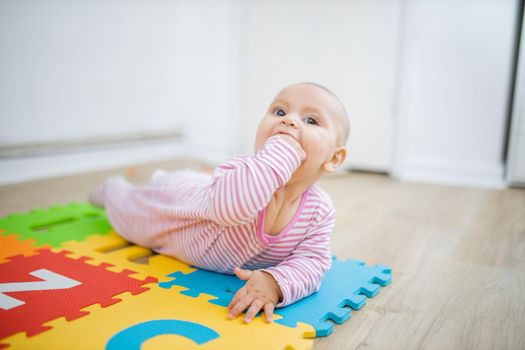 Adorable baby lying face down on colorful children mat with her fist in her mouth. Portrait of joyful baby playing on the floor. Happy babies having fun