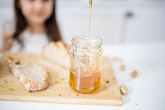 Picking honey from jar next to a slice of bread above wooden cutting board. Young girl at white table watching a honey jar.Sweet and light breakfast