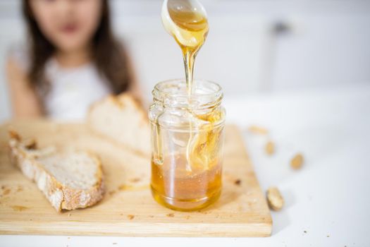 Male hand picking honey from jar next to a slice of bread above wooden cutting board. Young girl at white table watching a honey jar.Sweet and light breakfast