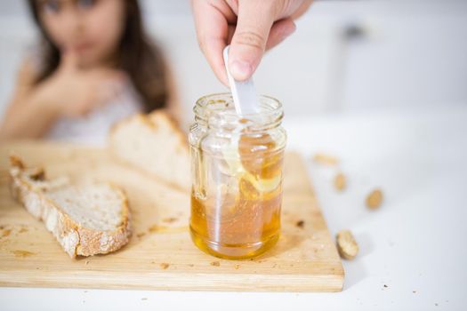 Male hand picking honey from jar next to a slice of bread above wooden cutting board. Young girl at white table watching a honey jar.Sweet and light breakfast