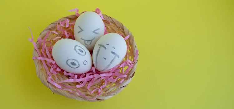 On the pink tinsel in the basket are three charming white eggs with faces on a yellow background. Easter Concept. Place for your text.