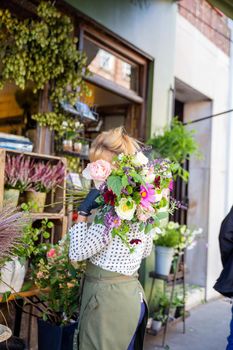 Brunette woman holding and covering her face behind bouquet of mixed flowers. Beautiful and colorful display of flowers outside flower shop. Houseplants and nature
