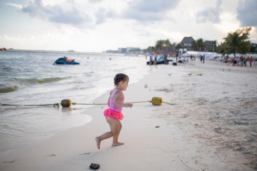 Adorable little girl joyfully running from the sea at the beach. Young child having fun on the beach under slightly cloudy sky. Tropical summer vacations