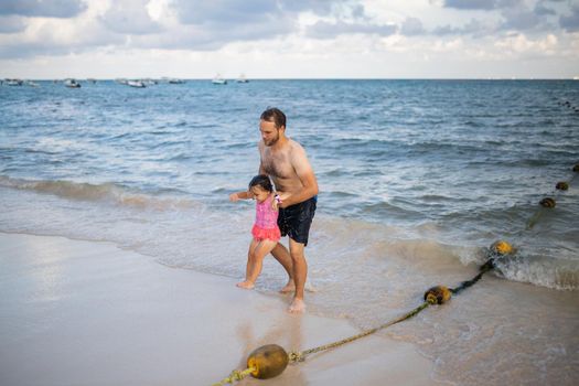 Happy father carrying adorable little daughter out of the water at beach. Man and cute young girl having fun on beach. Tropical summer vacations