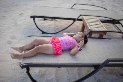 Happy and adorable little girl smiling and lying down on beach lounge chair. Cute young child calmly resting on the beach. Tropical summer vacations