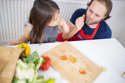 Happy little girl and her father at white table eating lemon slices. Man and young child cutting vegetables on wooden board. Daughter-father cooking together