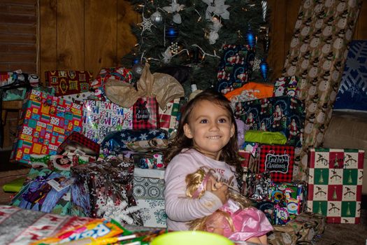 Happy adorable girl smiling and holding doll surrounded by presents and a Christmas tree. Joyful young child opening Christmas presents. Kids in winter celebrations