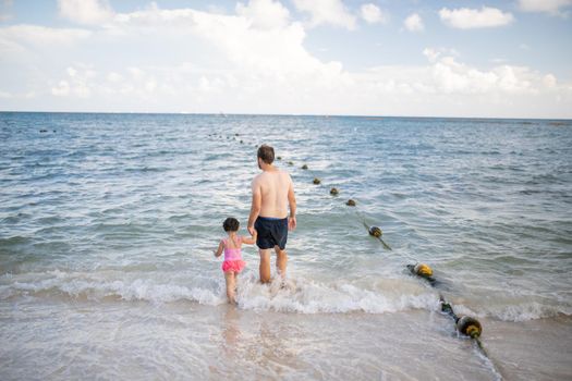 Father and young daughter holding hands as they walk on the beach and into the water. Man and little girl standing on the beach with beautiful sea in the distance. Tropical summer vacations