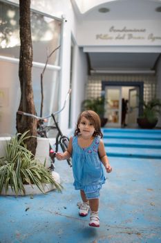 Adorable and vibrant little girl wearing blue dress playing at the entrance of white building. Portrait of cute young child having fun on sunny day. Tropical summer vacations
