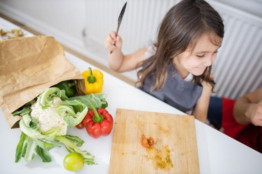 Happy little girl slicing cherry tomatoes on cutting board. Young child cutting vegetables on wooden board. Cauliflower, lemons, and peppers outside paper bag