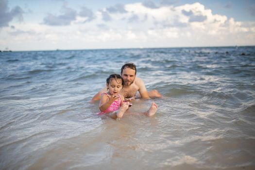 Happy father and young daughter joyfully playing in the water at the beach. Man and cute young child having fun in sea. Tropical summer vacations
