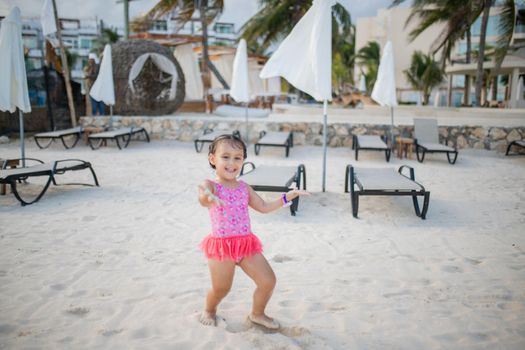 Happy adorable little girl smiling and playing with sand on the beach. Cute young child having fun with umbrellas and beach lounge chairs behind her. Tropical summer vacations