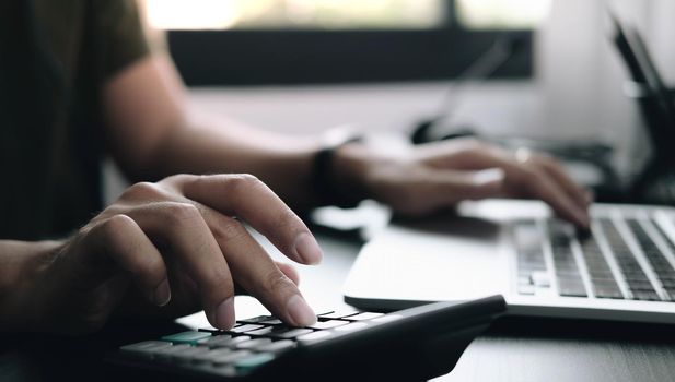 Close up Businessman using calculator and laptop for do math finance on wooden desk in office and business working background, tax, accounting, statistics and analytic research concept.