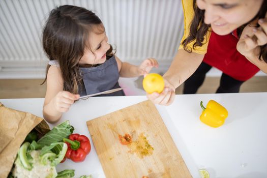 Happy little girl and her mother slicing lemons and tomatoes on cutting board. Smiling woman and young child at white talbe holding lemons. Daughter-mother cooking together