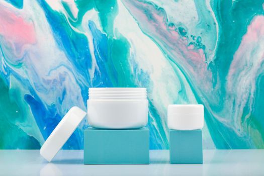 Face cream and lip balm on blue geometric podiums against blue marbled background. Concept of skin care and beauty products. High quality photo
