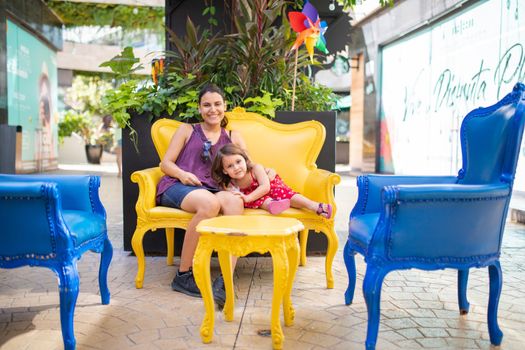 Happy mother and daughter sitting on yellow armchair in alley with plants behind them. Smiling woman and young girl resting on blue and yellow seats. Tropical summer vacations