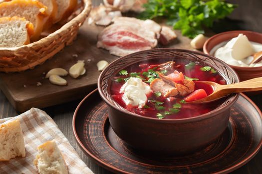 Freshly cooked hot homemade borscht - traditional dish of Russian and Ukrainian cuisine in earthenware dishes with bacon, bread, sour cream and garlic.