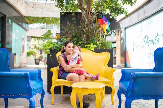 Happy mother and daughter sitting on yellow armchair in alley with plants behind them. Smiling woman and young girl resting on blue and yellow seats. Tropical summer vacations