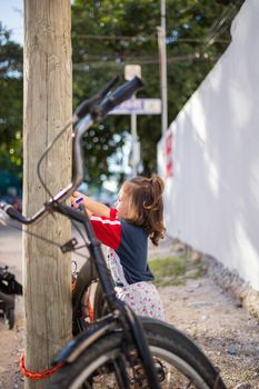 Adorable little girl playing next to bicycle that leans against wooden lamppost. Portrait of cute young child standing next to black bike in the street. Tropical summer vacations