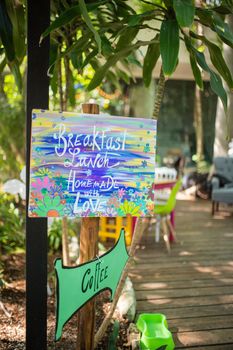 Colorful and flowered wooden sign for cafe with plants and trees as background. Sign for outdoors cozy and vintage-looking cafe. Rural cafeterias and restaurants