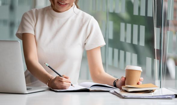 Cropped photo hand of woman writing making list taking notes in notepad working or learning online with laptop at home.