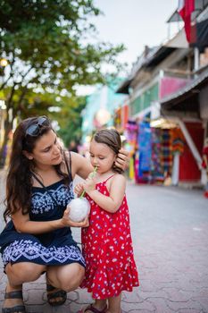 Beautiful woman and little girl drinking coconut water with straw in colorful alley. Portrait of happy mother kneeling next to cute young daughter. Tropical summer vacations