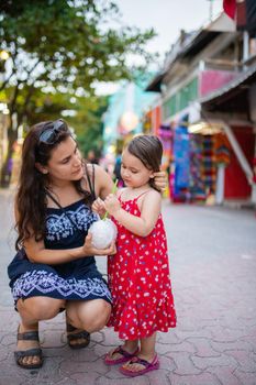 Beautiful woman and little girl drinking coconut water with straw in colorful alley. Portrait of happy mother kneeling next to cute young daughter. Tropical summer vacations