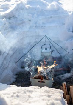 Over the fire hangs a pot in which to cook food. On a hook on a tripod, steam comes out of the pan. Winter Camping outdoor cooking